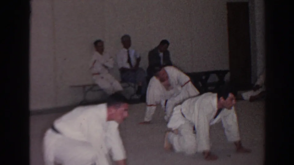 Adult trainees in white uniforms move low across the carpet while seated observers watch from the wall in the San Gabriel class footage.