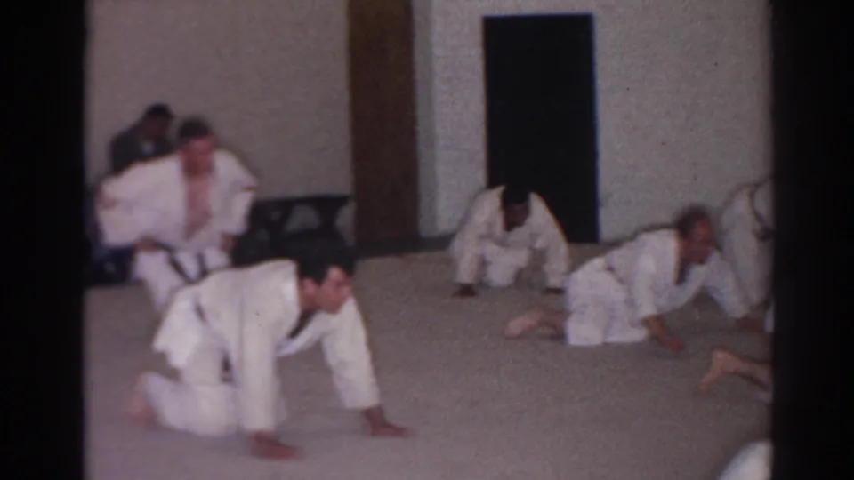Several adult men in white karate-style uniforms crawl or crouch across the floor in a simple indoor San Gabriel training space.