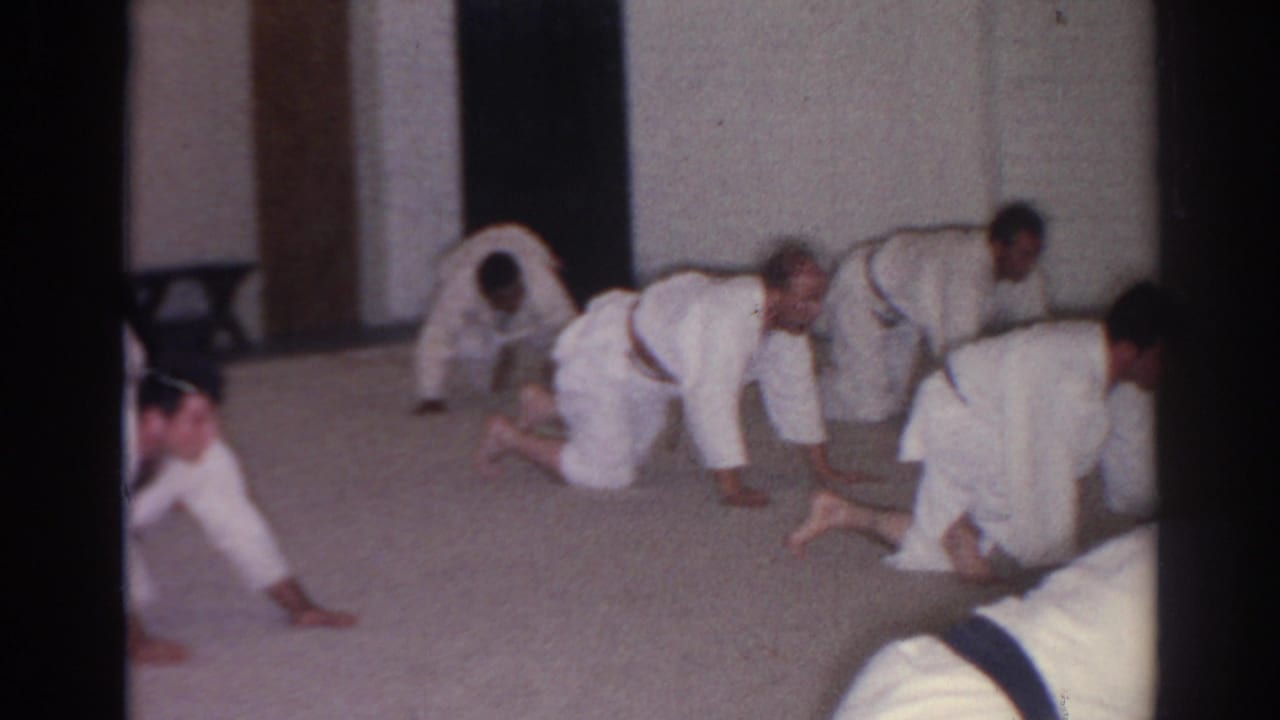 Men in white martial arts uniforms kneel and crawl across a carpeted indoor floor.