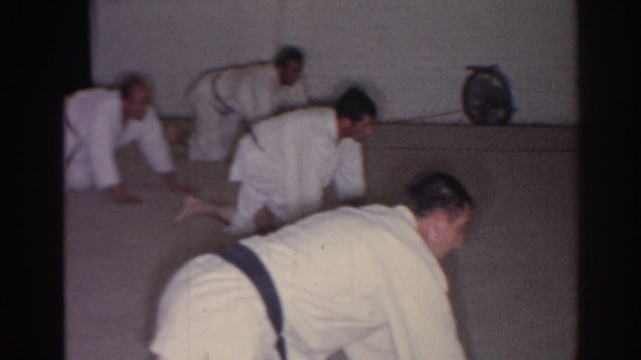 Several men in white uniforms crawl on a carpeted floor, with one figure close to camera.