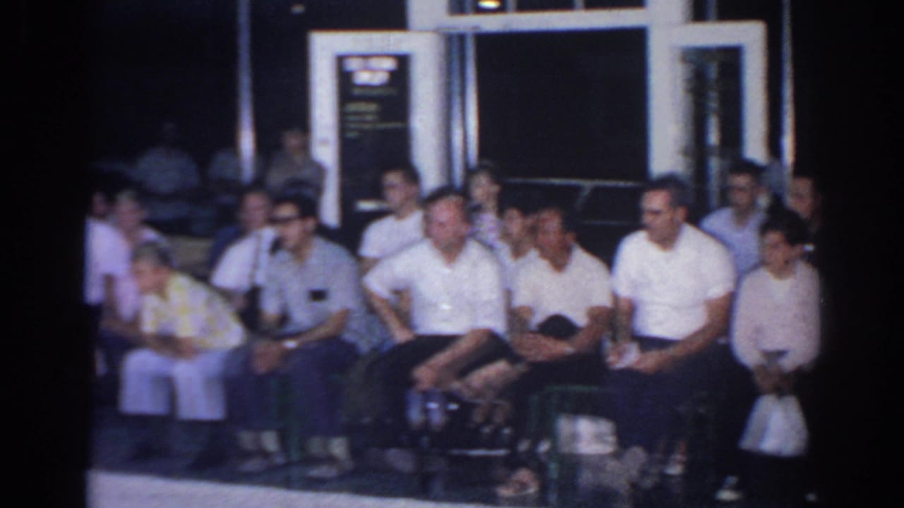 1966 San Gabriel home-movie frame showing seated spectators indoors, including men in white shirts and people on benches.