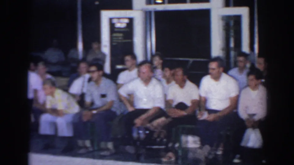 1966 San Gabriel home-movie frame showing seated spectators indoors, including men in white shirts and people on benches.
