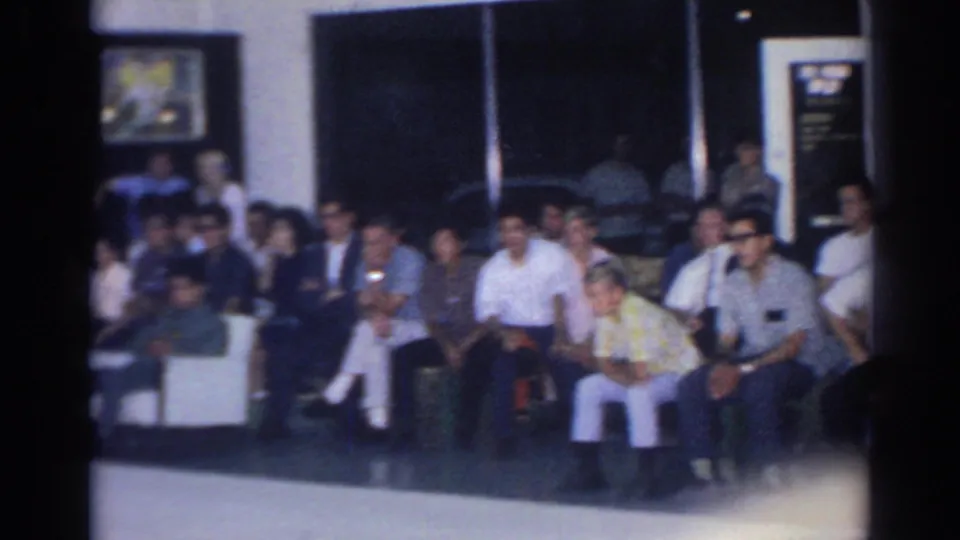1966 San Gabriel frame with a mixed group of spectators seated in rows along a dark interior wall.