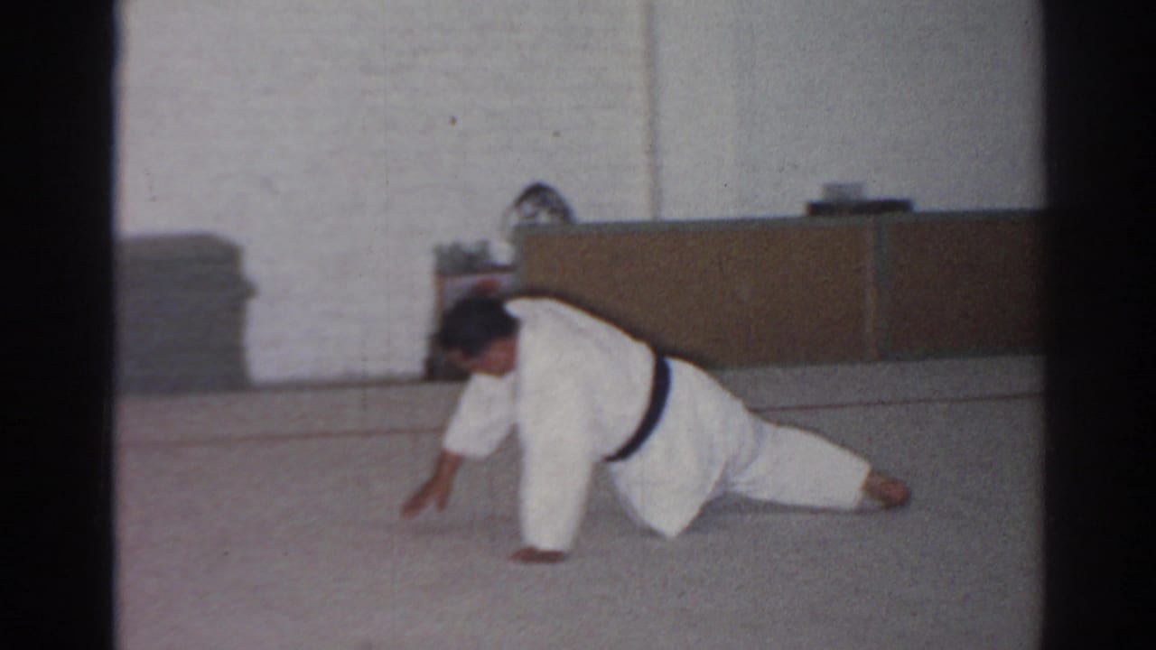 In 1966 San Gabriel footage, an adult man in a white gi and black belt stretches low on a gym mat near stacked mats and a cabinet.