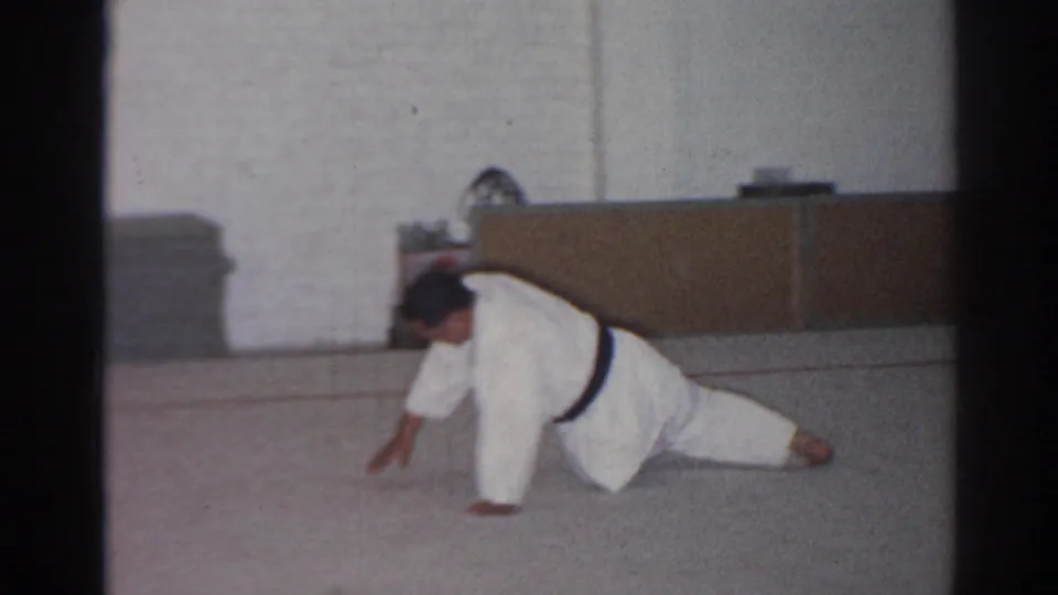 In 1966 San Gabriel footage, an adult man in a white gi and black belt stretches low on a gym mat near stacked mats and a cabinet.