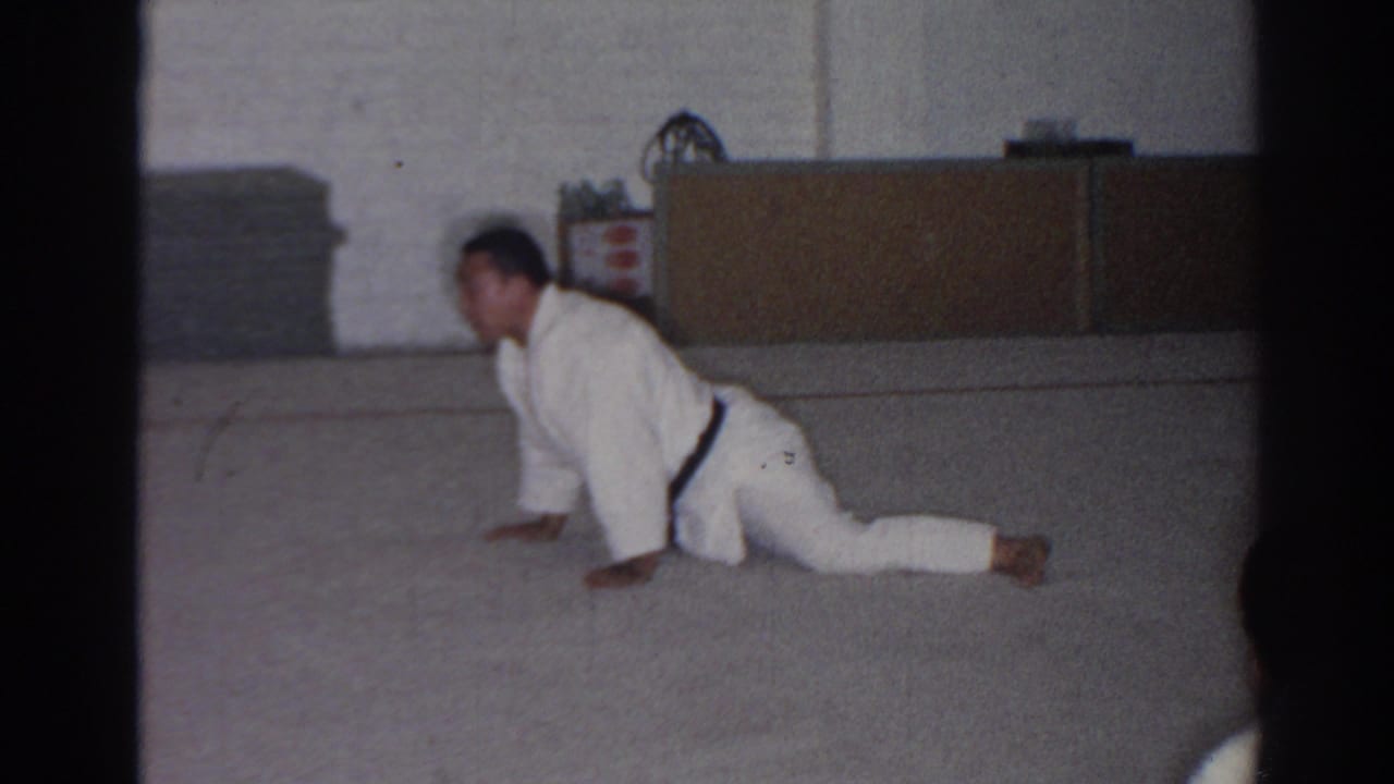 Man in a white gi holds a low push-up-like stretch on an indoor mat.