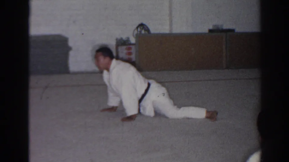 The man holds a low floor position in a white gi and black belt inside the San Gabriel gym setting shown in the 1966 home movie.