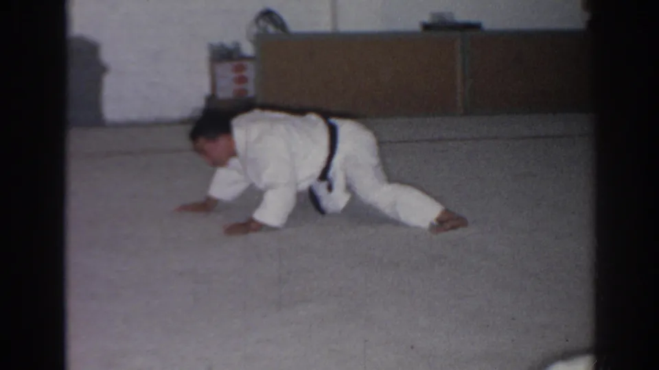 The adult man shifts through a low floor movement in the 1966 San Gabriel martial arts practice scene, with storage furniture behind him.