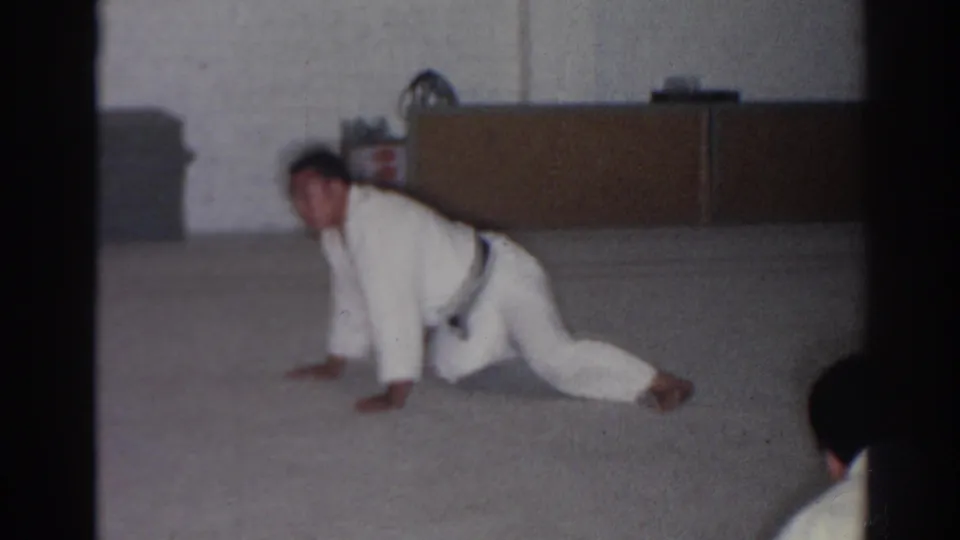 1966 San Gabriel home-movie frame showing a karate student in a white gi and dark belt holding a low floor lunge.