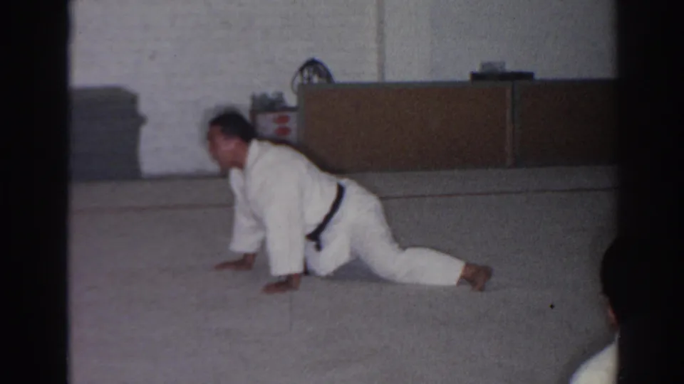 1966 San Gabriel frame with a karate student in a white gi stretching on the floor of a simple indoor training room.