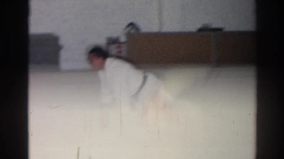 Soft 8mm frame from San Gabriel showing a karate student bending forward during a floor stretch in a plain indoor room.