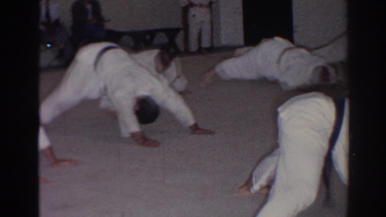 Men in white judo uniforms perform push-ups on an indoor carpeted floor.