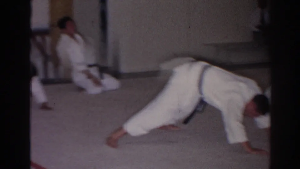A judo student in a white gi pushes forward near the camera while another uniformed participant kneels in the room.