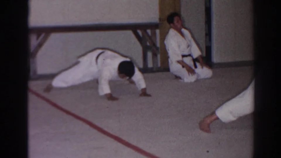 A man in a white judo gi holds a low push-up position as another uniformed participant kneels near the wall in the training room.