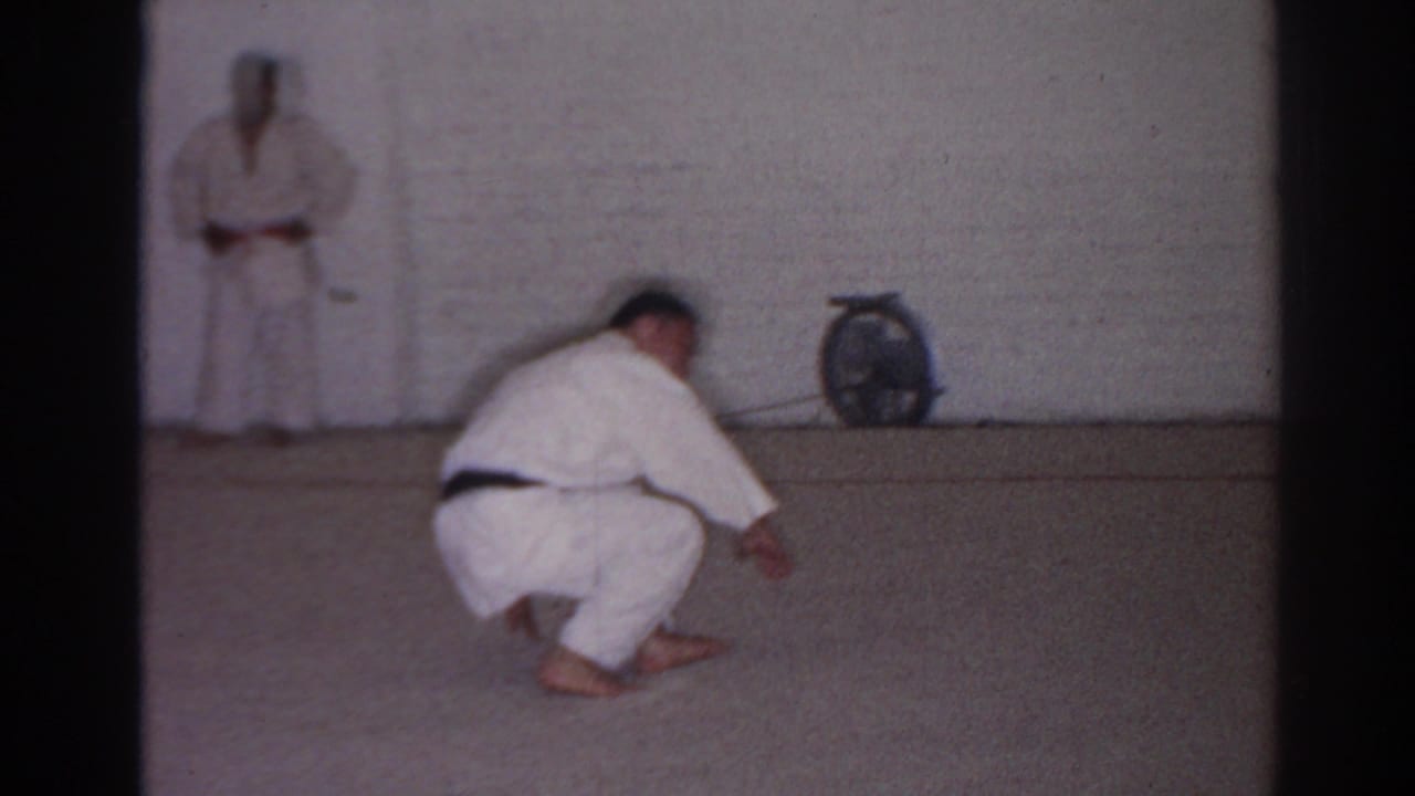 Martial arts student in a white gi crouches on the dojo floor while another person stands near the wall.
