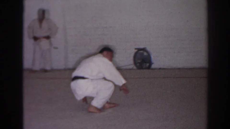 1966 San Gabriel home-movie frame showing a martial arts student in a white gi crouching before a roll in a simple dojo.