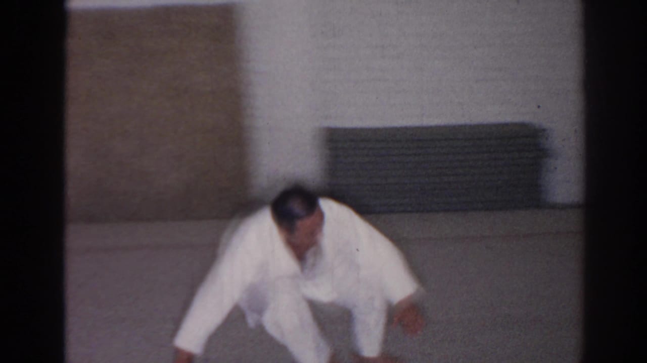 Student in a white gi moves forward close to the floor in a dojo interior.