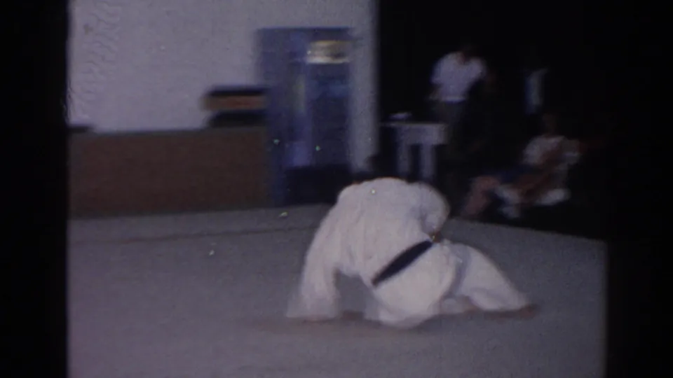 1966 San Gabriel dojo frame with a student low on the floor after a roll, with spectators seated along the wall.