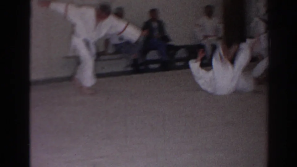 1966 San Gabriel home-movie frame showing martial arts students during a tumbling or rolling demonstration with spectators behind them.
