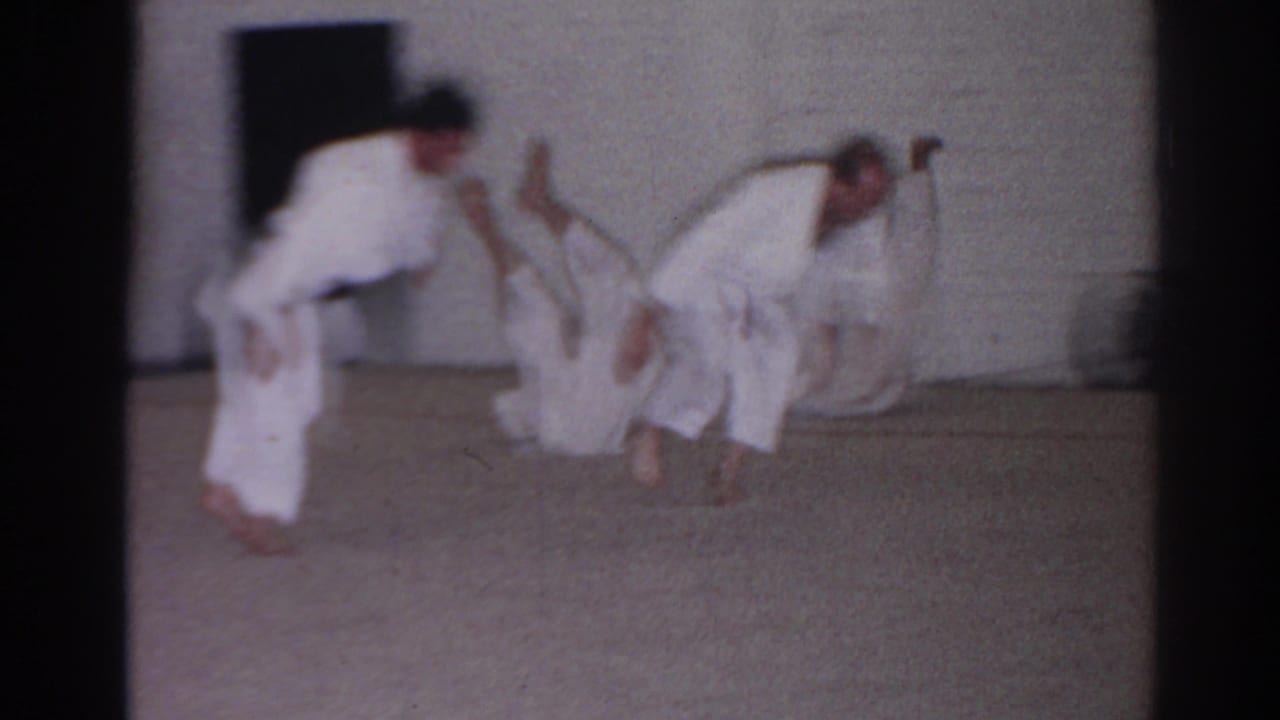 Students in white martial arts uniforms practice rolling movements on a carpeted room floor.
