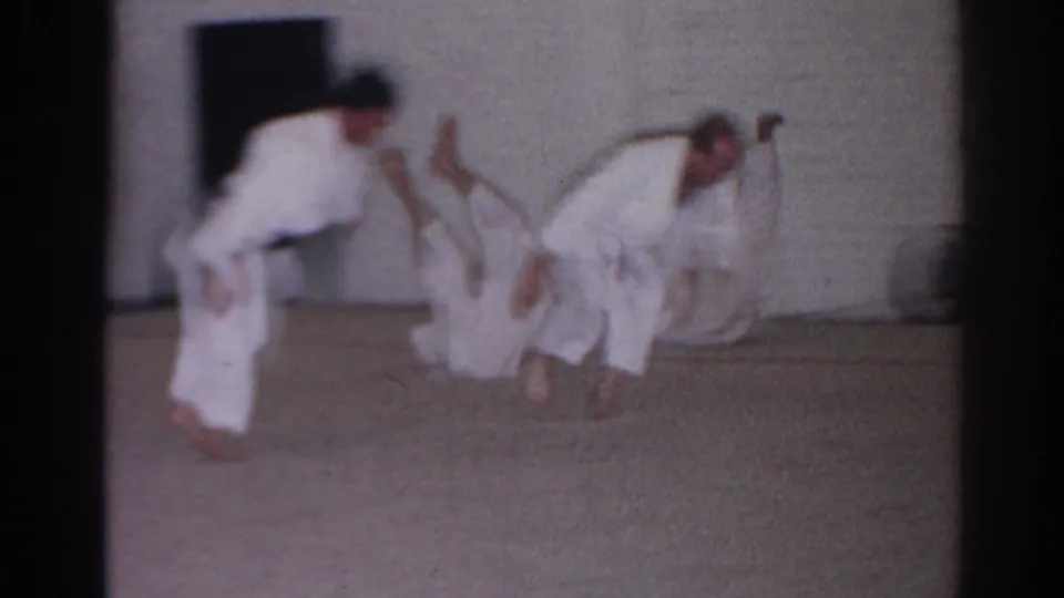 1966 San Gabriel home-movie frame showing martial arts students in white gi practicing rolls in an indoor class space.