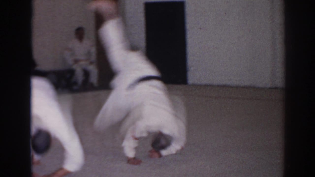 1966 San Gabriel frame with a student in a white gi rolling forward as classmates observe from the room edge.
