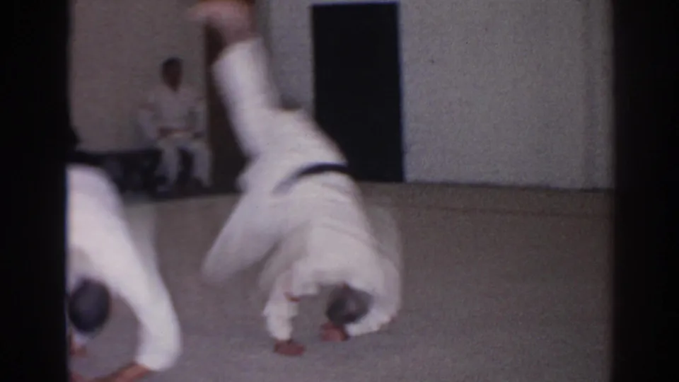 1966 San Gabriel frame with a student in a white gi rolling forward as classmates observe from the room edge.