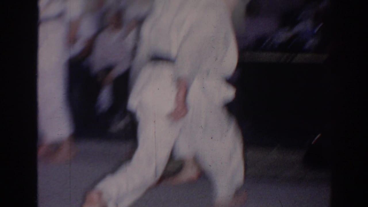 Soft 8mm frame from a 1966 San Gabriel karate practice, showing white gi-clad students moving across indoor mats.