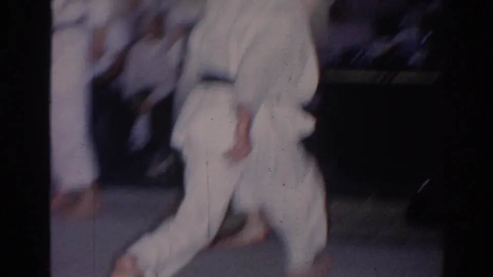 Soft 8mm frame from a 1966 San Gabriel karate practice, showing white gi-clad students moving across indoor mats.