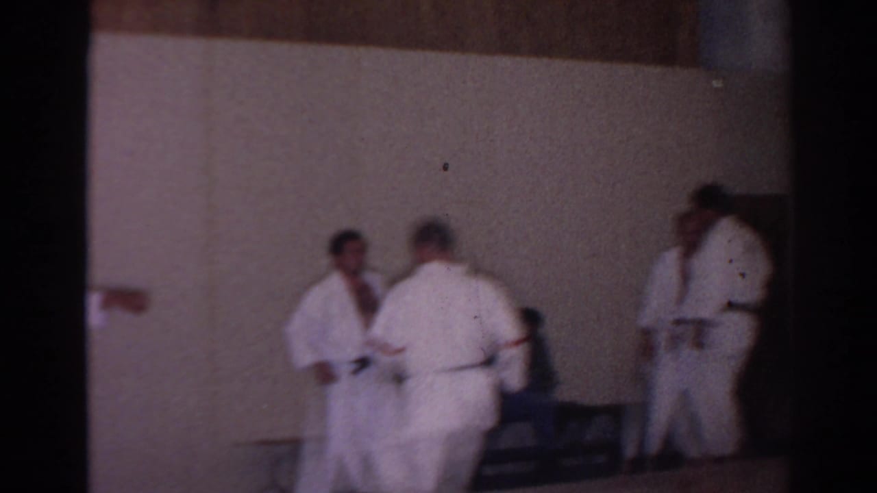 Adult karate students in white gi stand and confer near chairs in a plain San Gabriel training room, 1966.