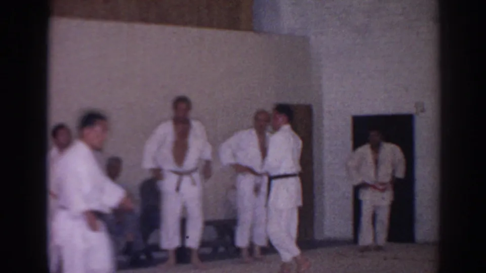 A 1966 home-movie frame shows karate students in gi standing on mats as another participant passes close to the camera.