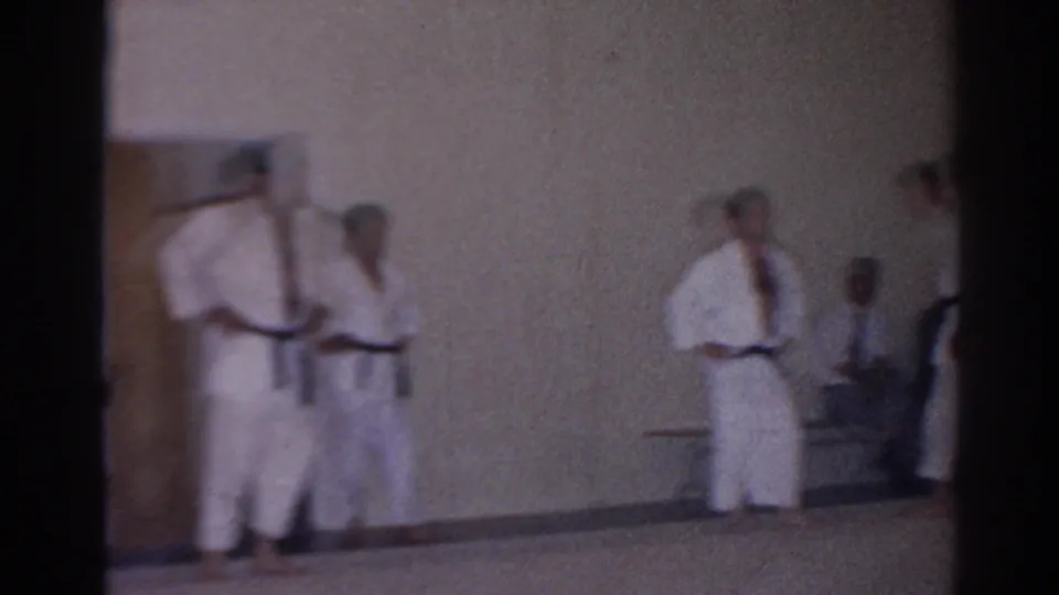 Wide, soft 8mm view of adult karate students in white gi practicing or waiting in an indoor San Gabriel class setting.