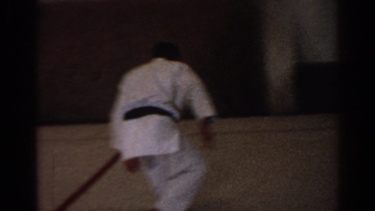 1966 San Gabriel home-movie frame showing a martial arts practitioner in a white gi moving in a simple indoor training room.