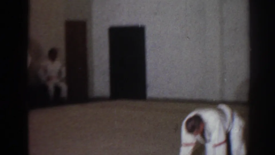 1966 indoor training frame showing a crouched martial arts participant and another white-uniformed figure near dark doorways.
