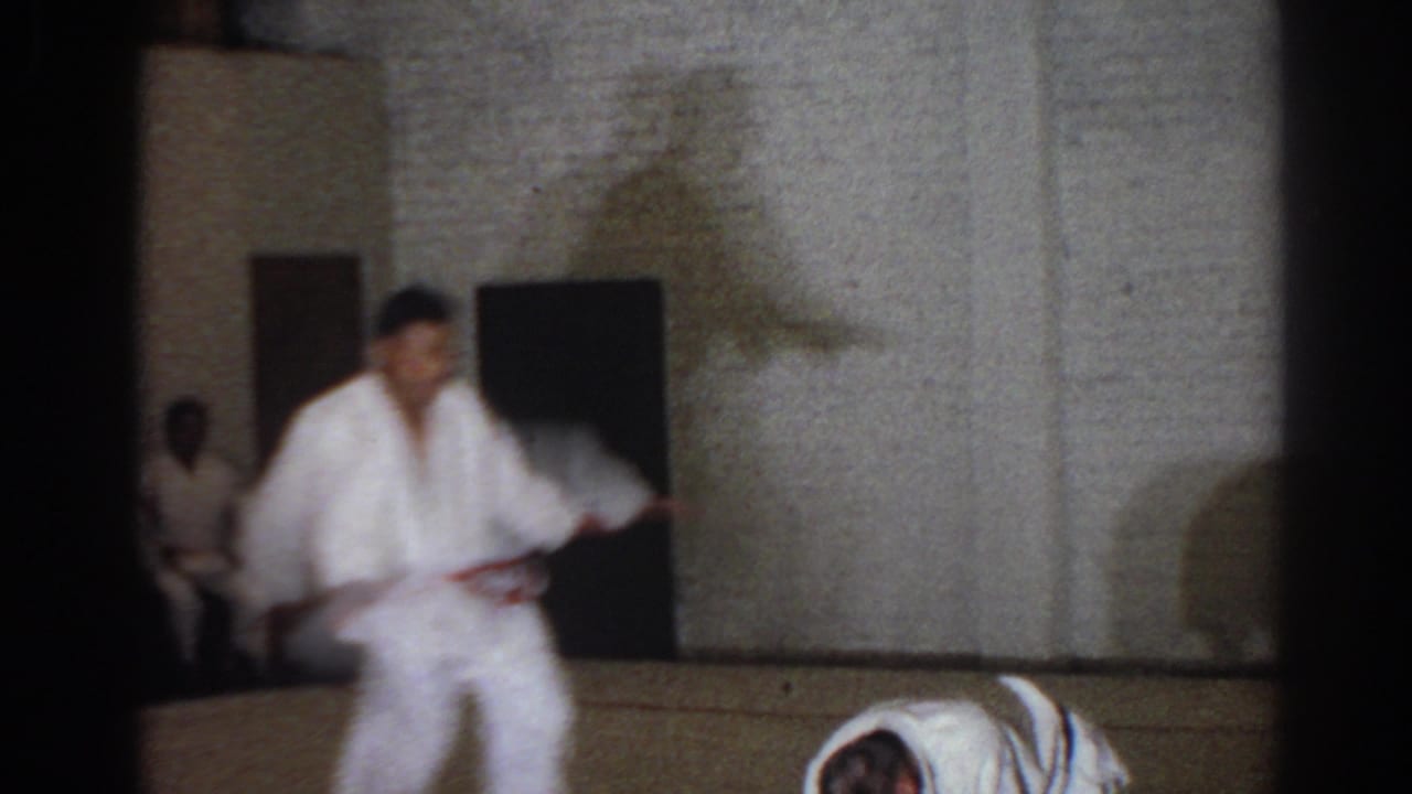 A man in a white uniform steps across a mat while another participant crouches nearby.