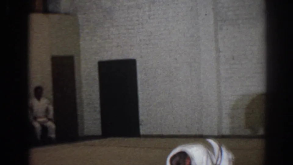 1966 San Gabriel home-movie frame showing a quiet dojo wall, seated observer, and a uniformed participant near the floor.