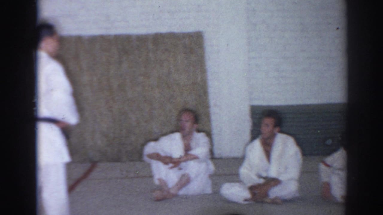 Young men in white gi sit on a mat while an instructor stands at the edge.