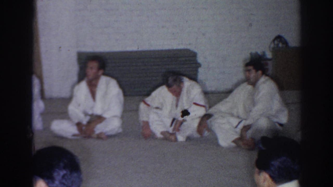Martial arts students in white gi sit along a mat near stacked pads.