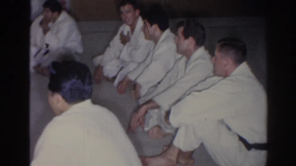 Young men in gi sit in a loose circle on a mat in a San Gabriel martial arts class, filmed in 1966 on 8mm.