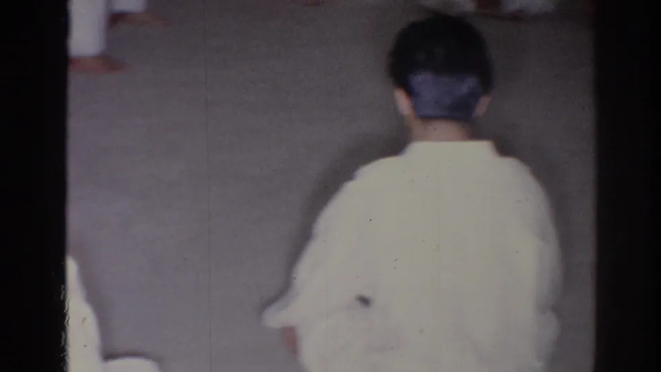 1966 San Gabriel frame of a martial arts practitioner in a white gi viewed from behind on a gym-like floor.