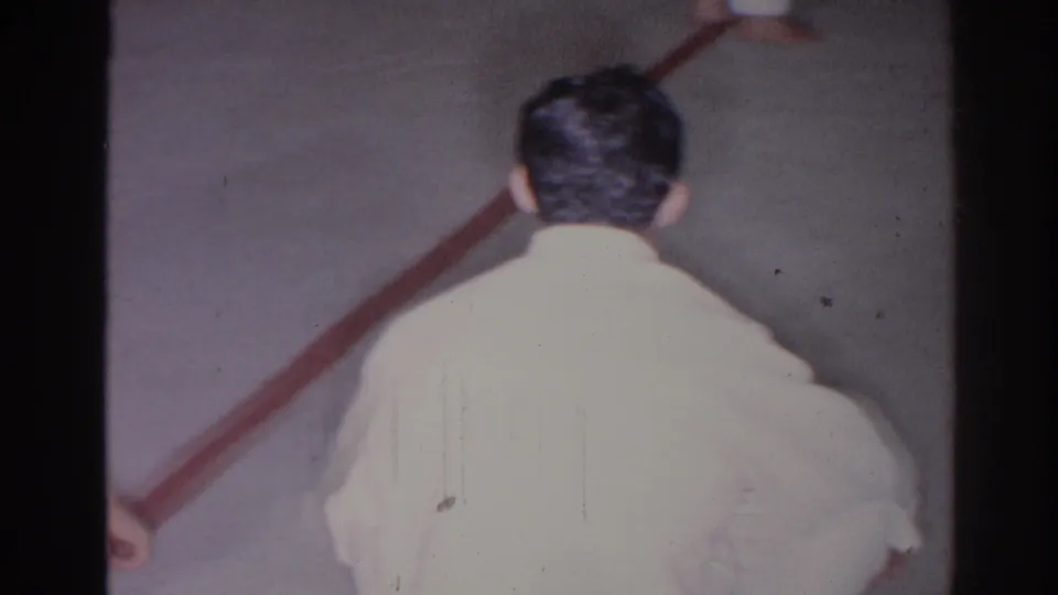 1966 San Gabriel home-movie still showing a white-uniformed martial arts participant from behind near a red line on the floor.
