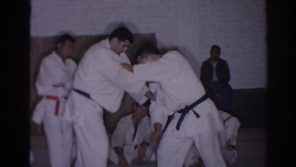 1966 San Gabriel gym scene with two judo practitioners in white gi demonstrating a technique before seated students and onlookers.