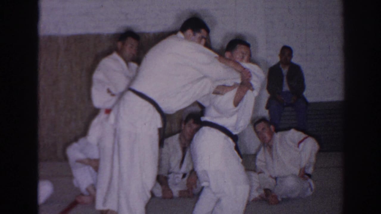 In a 1966 San Gabriel gym, two black-belt judo practitioners demonstrate a close standing hold while students sit along the wall.