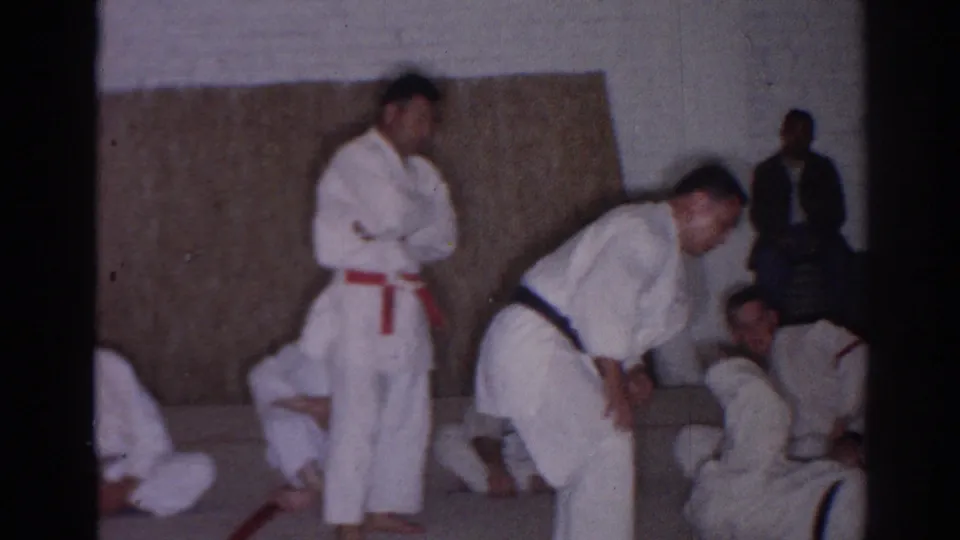 1966 San Gabriel judo demonstration frame showing one practitioner bent forward, a red-belt student standing, and others seated along the gym wall.