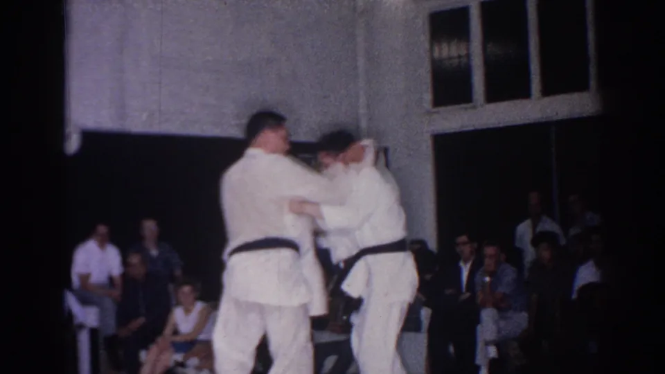 1966 San Gabriel gym footage of two judo practitioners grappling before a small audience seated near windows and a dark doorway.
