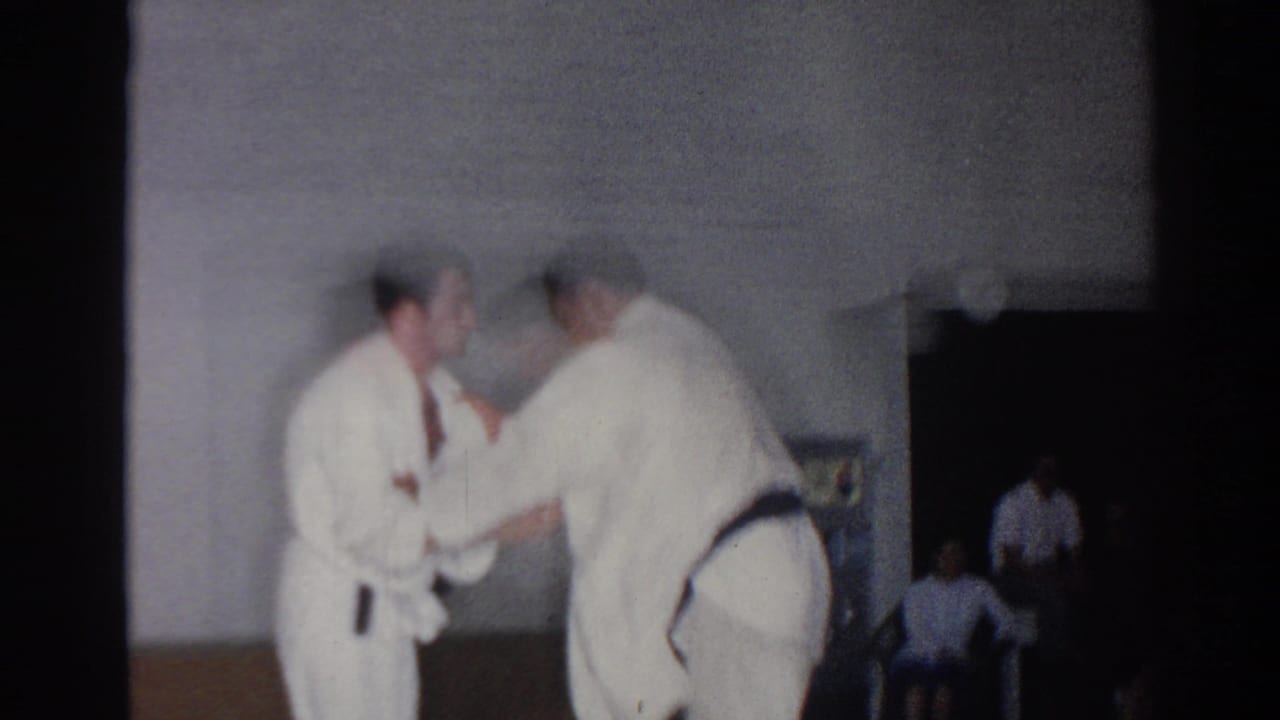Two men in white judo uniforms practice a grappling technique in a gym.