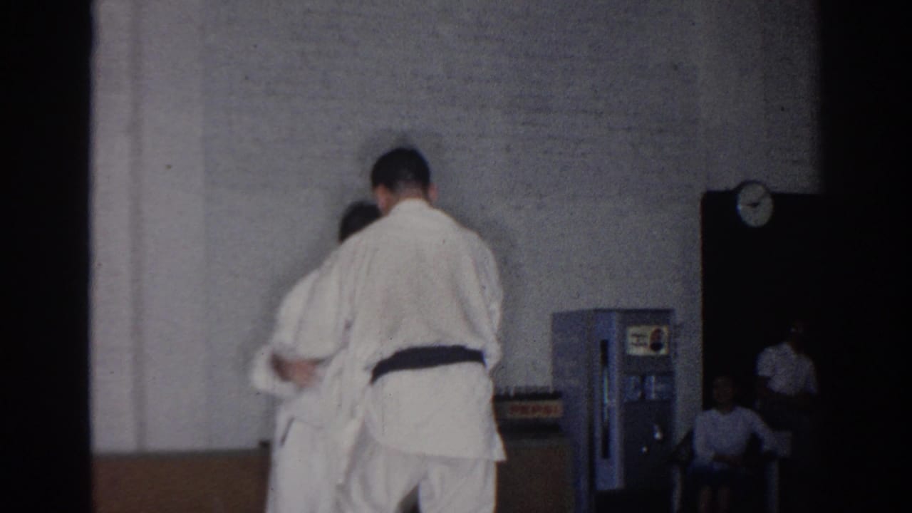 A judo practitioner in a white gi holds another person near a wall clock and vending machine.