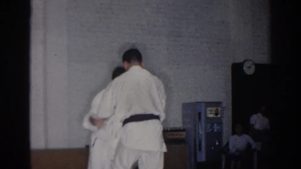 A 1966 San Gabriel judo practice moment shows one man in a white gi holding a partner near a vending machine and wall clock.