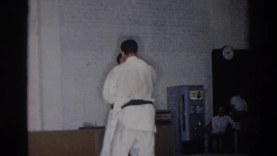 Two men in judo uniforms continue a close drill in a San Gabriel gym, with seated spectators visible near the doorway.