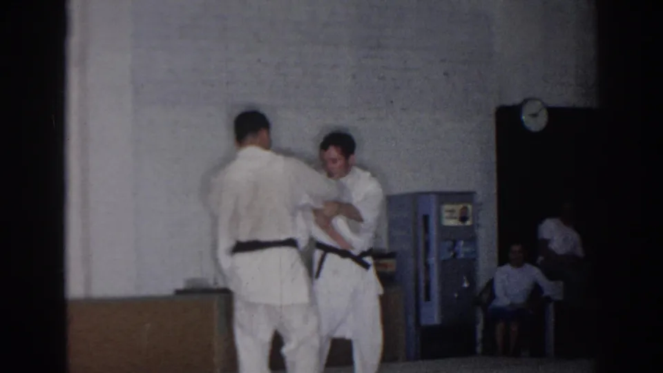 At the end of the 1966 San Gabriel clip, two judo practitioners reset or adjust grips beside a vending machine and seated onlookers.